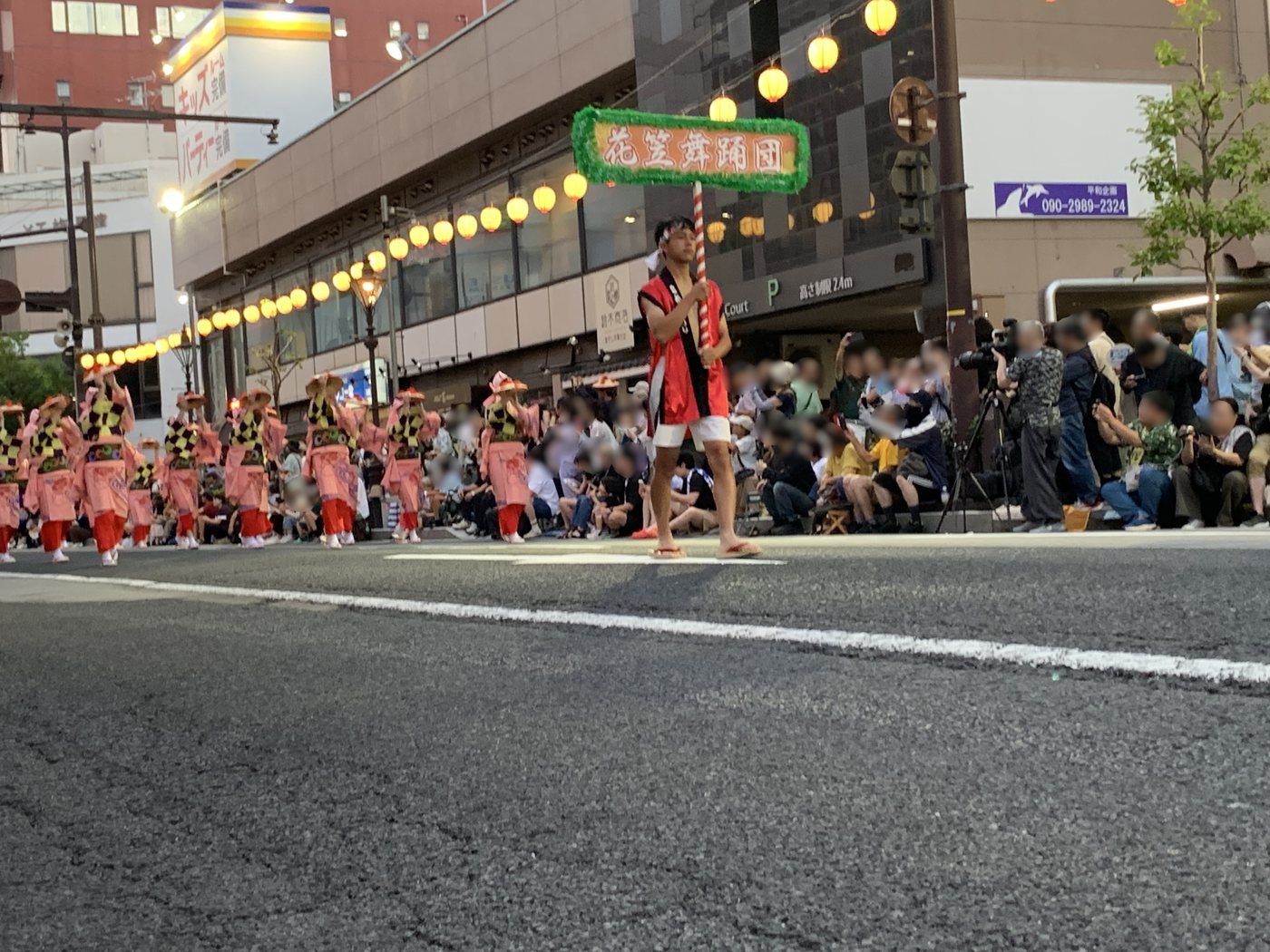日が落ちた沿道。歩道に観客が座り、提灯の下を踊り手が進む